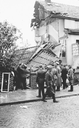 Bombentreffer. Eine Seitenwand eingestürzt. Zivilisten und Uniformierte auf der Strasse.